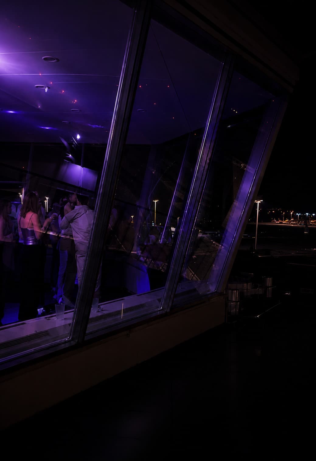 Gäste an der Bar der Lounge mit Blick über das nächtliche Vorfeld des Flughafens Zürich